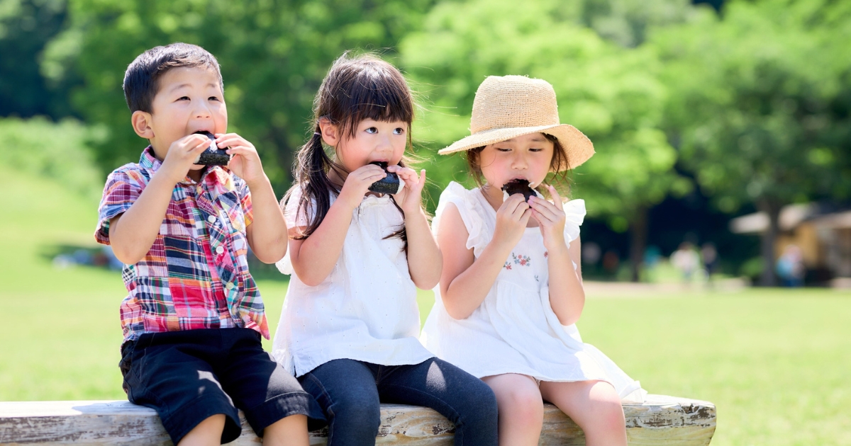 3人の子どもがベンチに座っておにぎりを食べる様子
