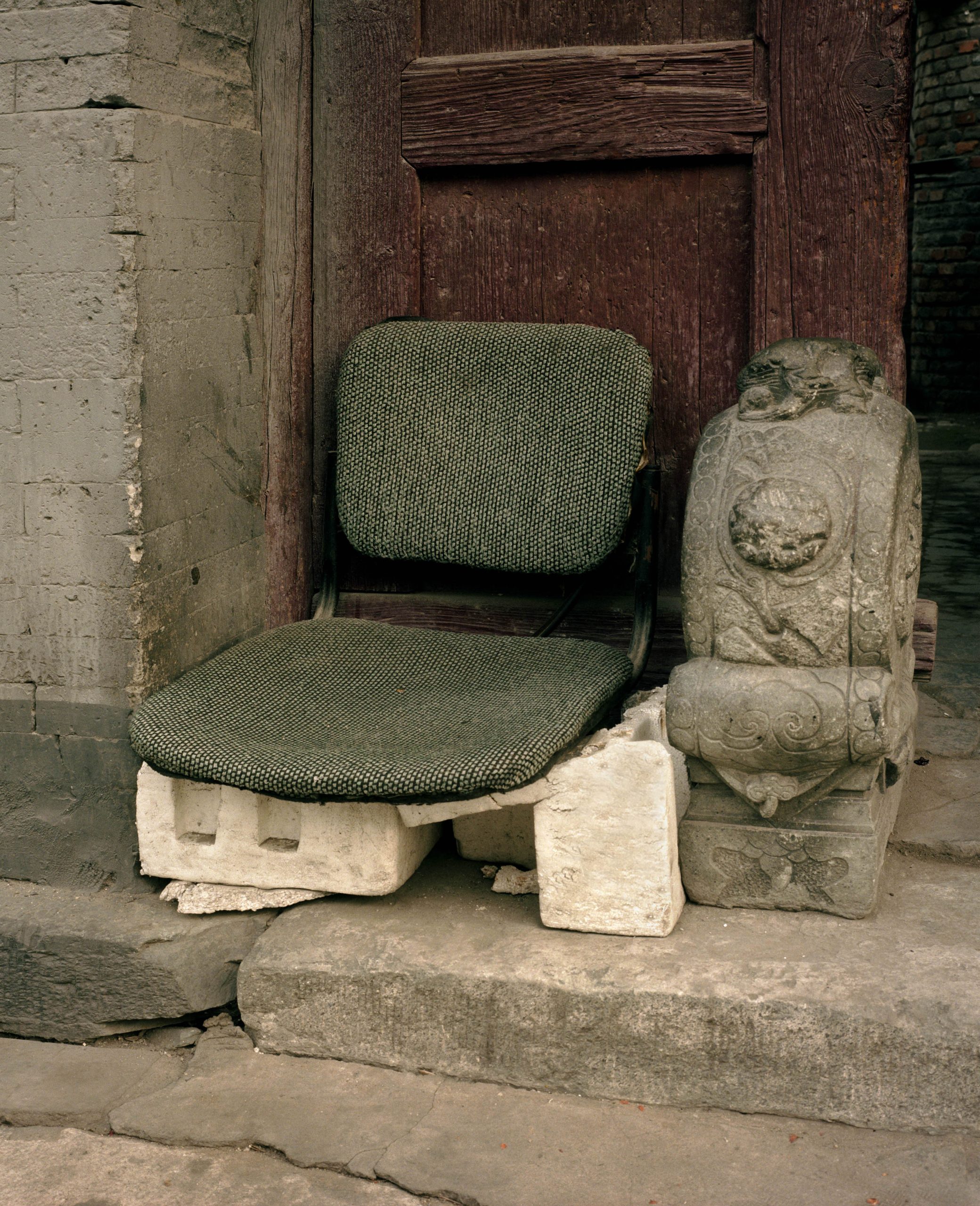 Michael Wolf, Bastard Chairs. A green upholstered seat stands low on blocks at a doorway.