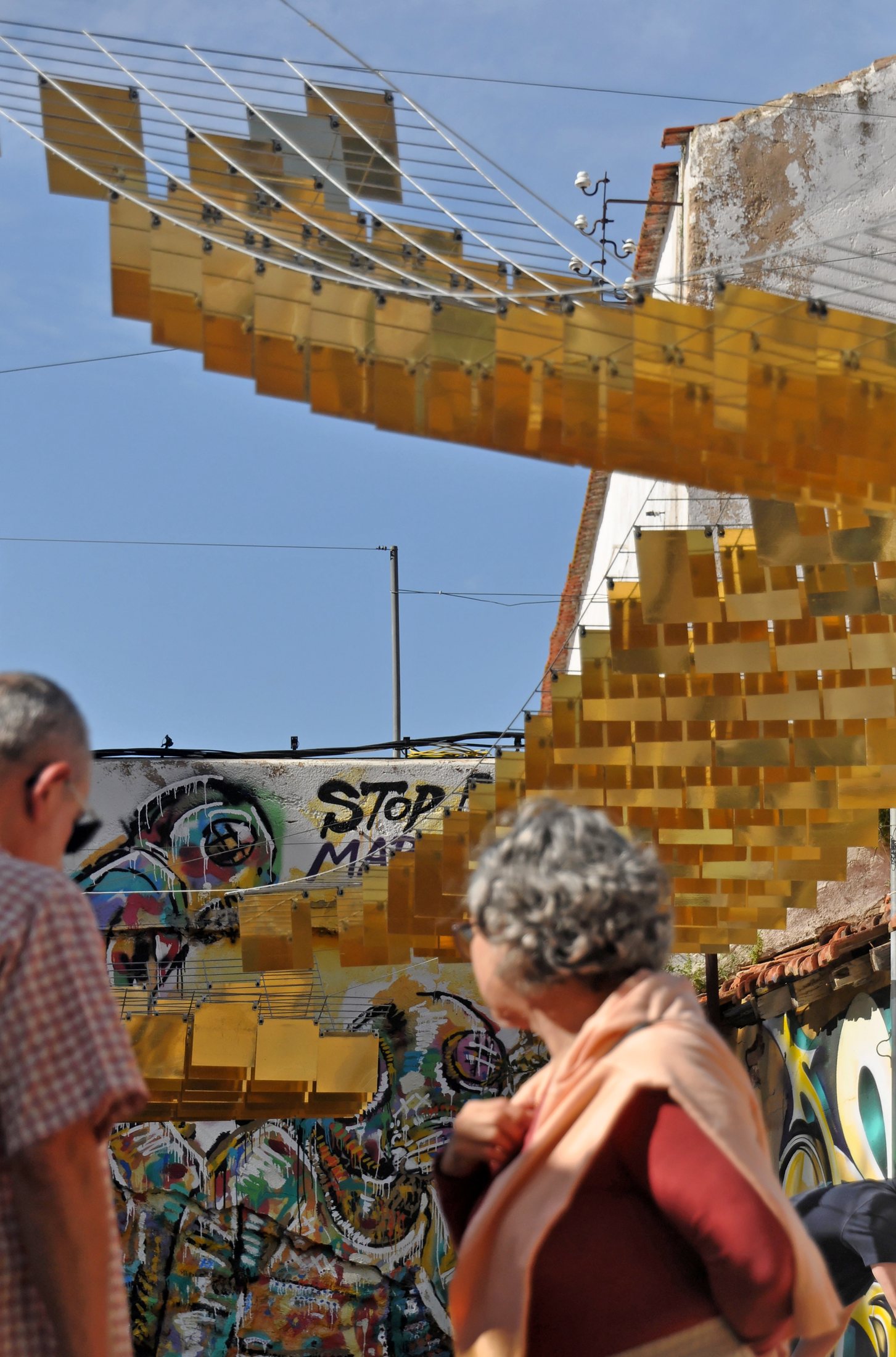 Visitors standing beneath the suspended golden pieces of A-L-M-A-D-R-A-V-A